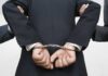 Person handcuffed in suit, escorted by two others.