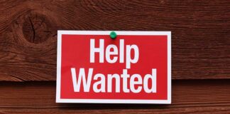 A red and white 'Help Wanted' sign pinned to a wooden surface