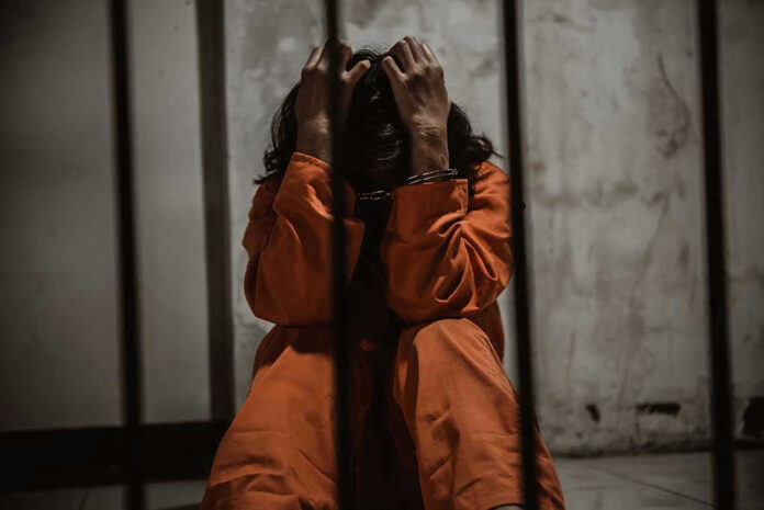 Person in orange jumpsuit sitting behind bars, handcuffed.
