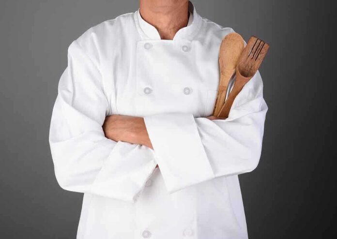 A chef in a white uniform holding wooden cooking utensils with crossed arms