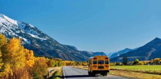 A yellow school bus driving on a rural road surrounded by autumn trees and mountains