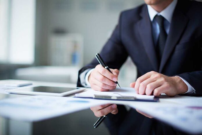 A man in a suit writing notes at a desk with documents and a tablet