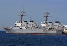 Warplanes Fly, Diplomacy Waits Three gray naval destroyers docked side by side at a pier
