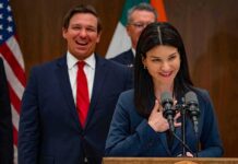 Ron DeSantis smiling behind a woman speaking at a podium