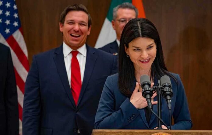Ron DeSantis smiling behind a woman speaking at a podium