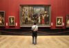 Lawsuits Target Alleged Nazi-Looted Art A person standing in an art gallery, observing a large historical painting surrounded by other artworks