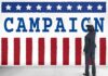 Trump Repeats Anti-Socialism Pledge Man in suit writing on large campaign banner with stars and stripes